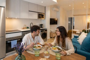 A modern kitchen with stainless steel appliances, a dining table with two people eating, and a living area with a blue sofa.