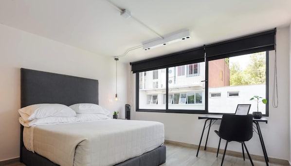 A modern bedroom with a large bed, a desk, and a window with a view of buildings.