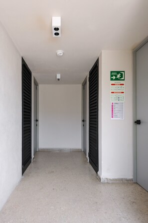 A hallway with a ceiling-mounted smoke detector and a green emergency exit sign.