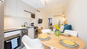 Een moderne keuken met roestvrijstalen apparatuur, een eetgedeelte met een houten tafel gedekt voor twee personen, en een zithoek met een bank en decoratieve kussens.