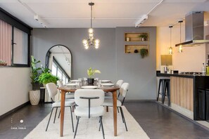 Un comedor moderno con una mesa redonda, sillas blancas y una araña. Hay un espejo grande en la pared, una planta en maceta y una zona de cocina visible al fondo.