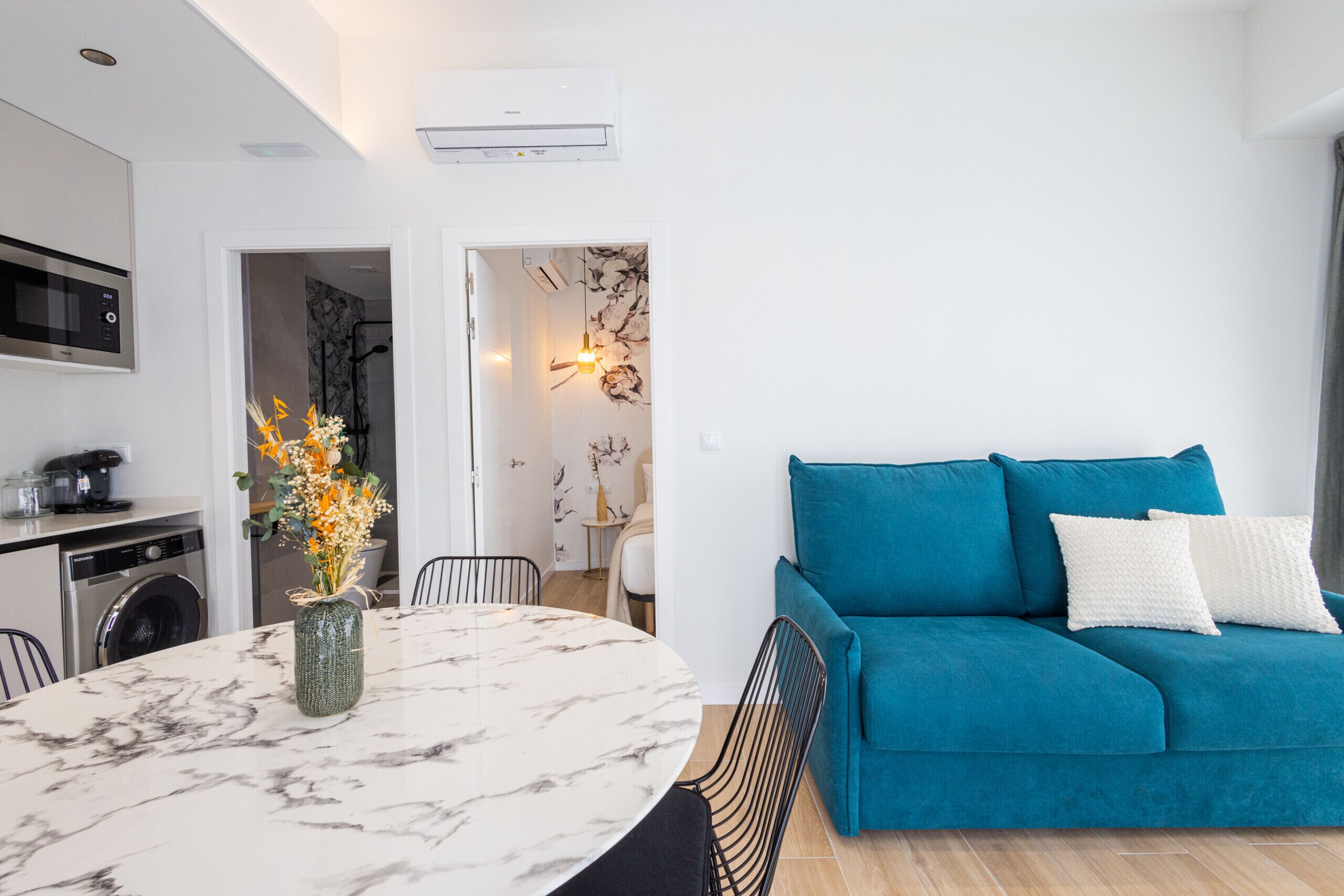 Un salon moderne avec un canapé de couleur turquoise, une table à manger en marbre et un coin cuisine équipé d’un lave-linge.