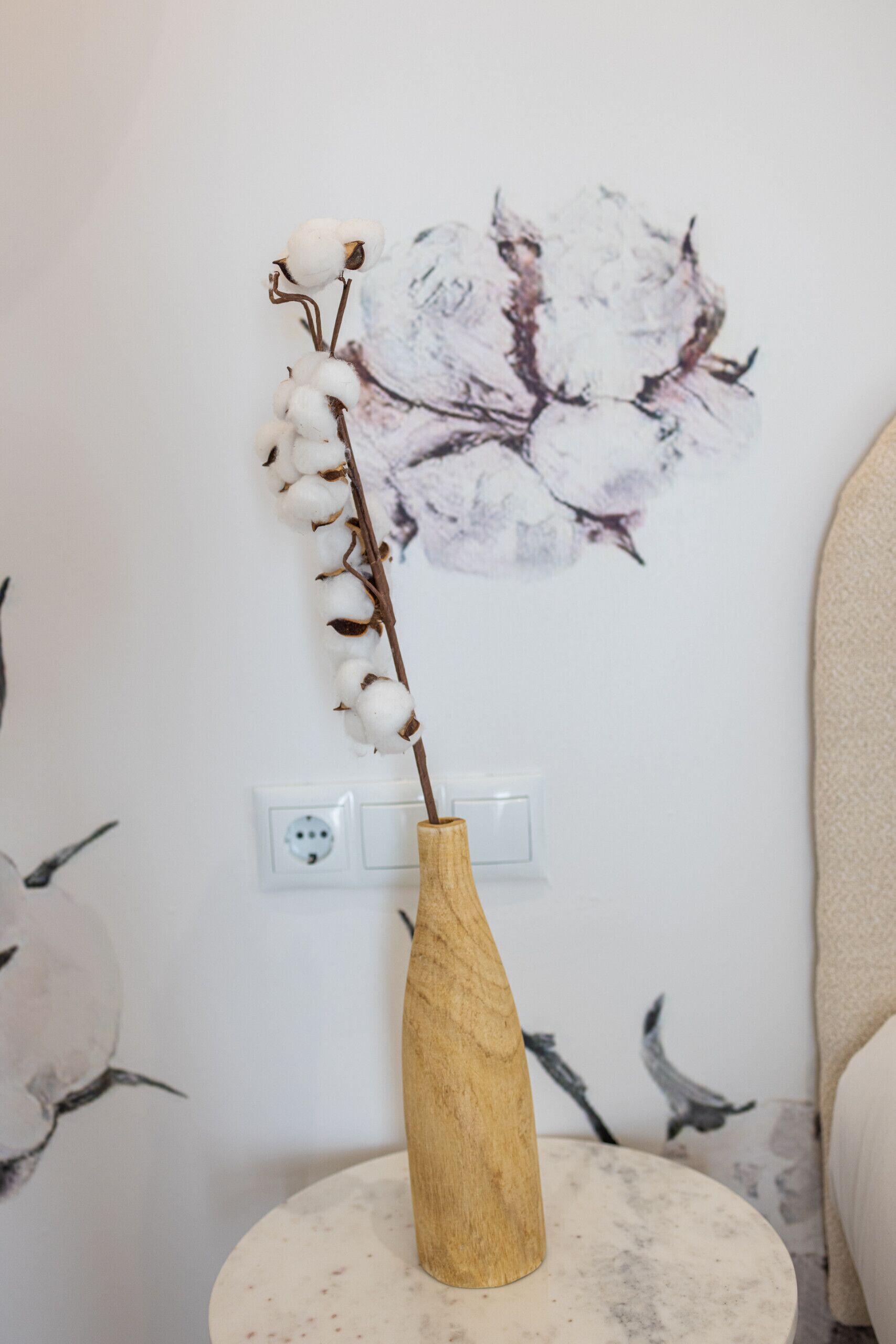 A wooden vase with cotton branches on a round marble table against a wall with a power outlet and a switch.