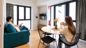 A modern living room with a sofa, a dining table with chairs, a TV mounted on the wall, and large windows with curtains. - Turia Park Loft IV | 4 Guests | AC and WiFi | Near ZOO (Valencia)