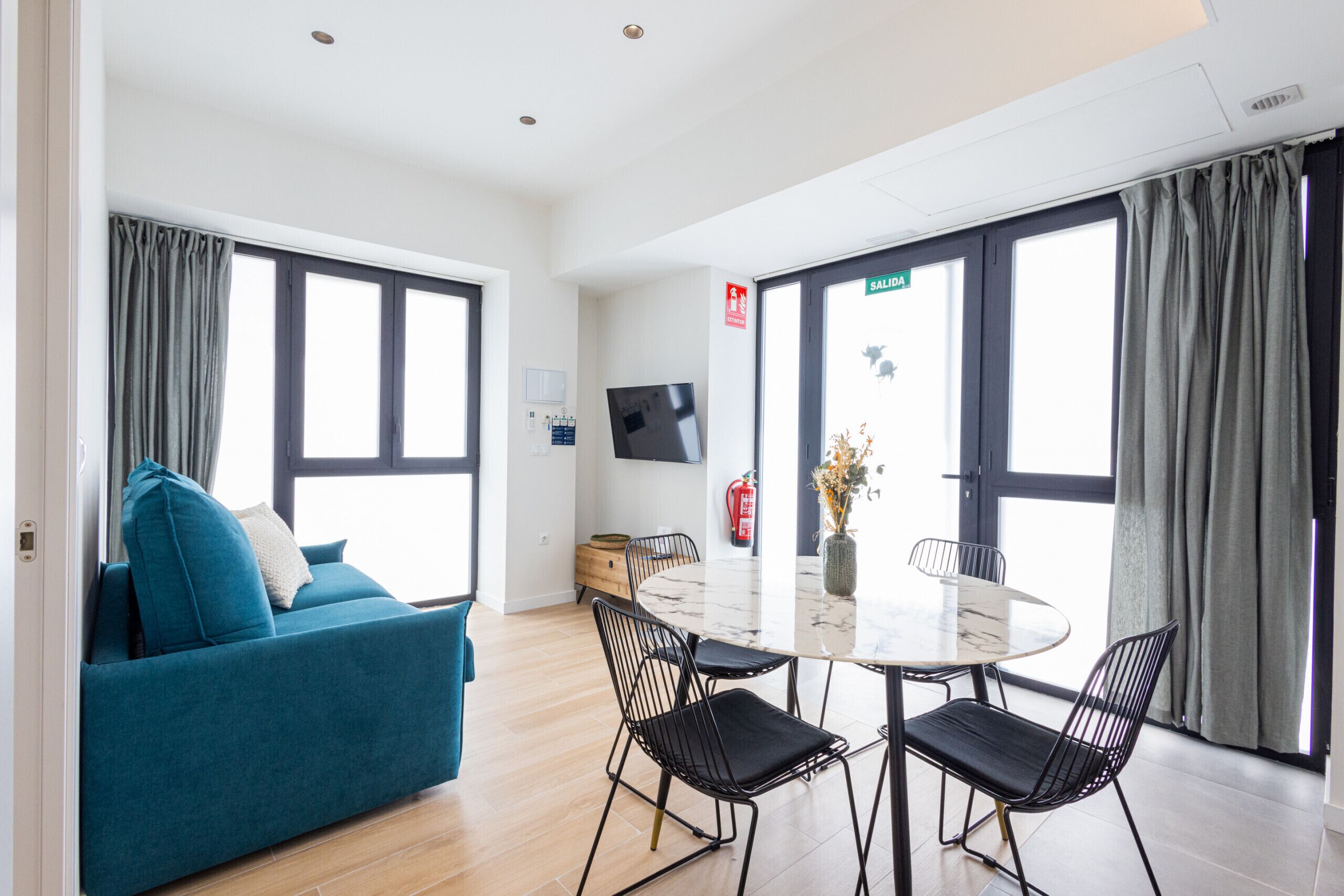 Una habitación moderna con sofá, una mesa de comedor con sillas, televisor de pantalla plana y ventanas grandes con cortinas.