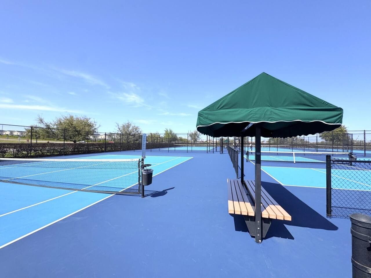 An outdoor tennis court with a green pavilion, benches, and a trash can.
