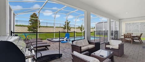 Un espacio de patio cubierto con mobiliario exterior, una parrilla de barbacoa y vistas a una piscina y follaje.