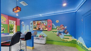 Een kamer met een muurschildering in Nintendo-thema, een Nintendo Switch-console en een barruimte met stoelen.