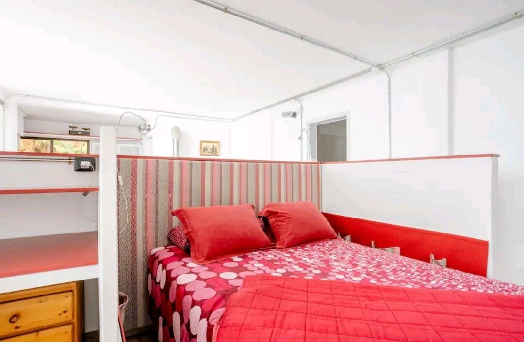 Een stapelbed met rode beddengoed en een gestreept hoofdeinde, een klein raam en een houten dressoir.