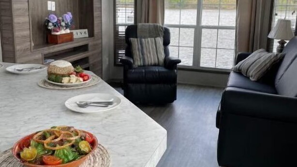 A modern living room with a marble dining table, a black leather armchair, and a black sofa. There is a ceiling fan, a large window with a view of a body of water, and a wooden shelf with decorative items.