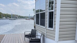 A wooden deck with two chairs and a table, overlooking a body of water with boats and a bridge in the distance.