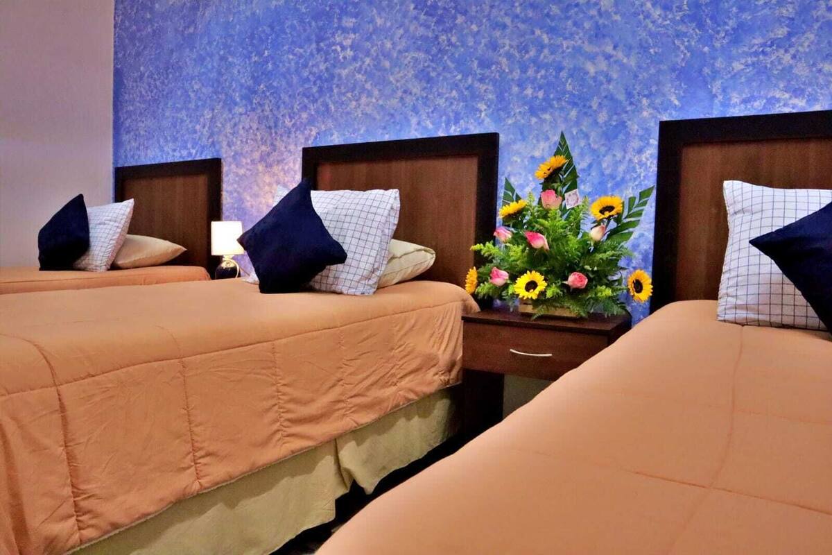 Habitación de hotel con tres camas, cada una con cabecera y varias almohadas. Se observa un mesita de noche con lámpara y un arreglo floral entre las camas.