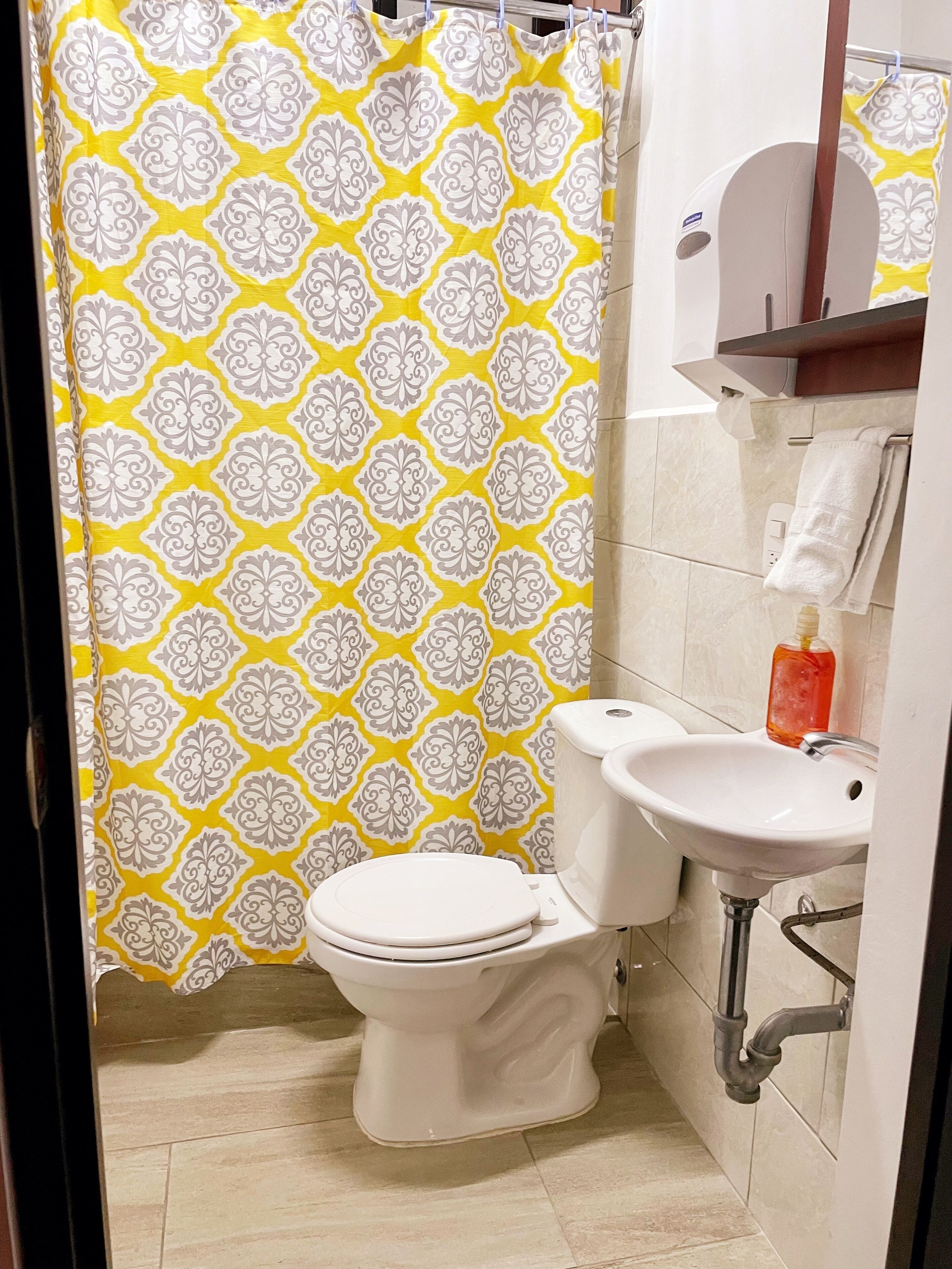 Un baño con cortina de ducha de diseño amarillo y blanco, un inodoro blanco, un lavabo blanco y un dispensador de jabón.