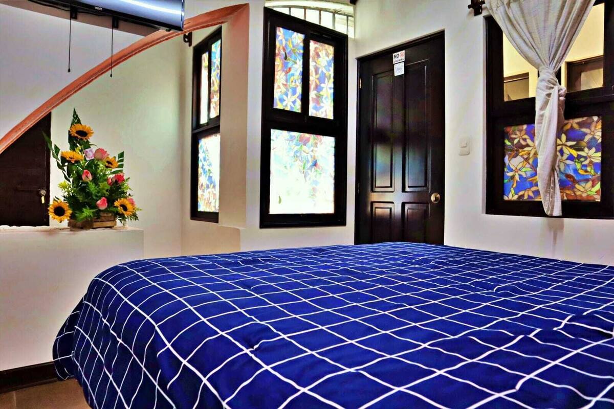 Un dormitorio con una cama grande, un ventanal con vidrieras, una puerta con letrero de 'No Fumar' y un arreglo floral sobre una mesa.