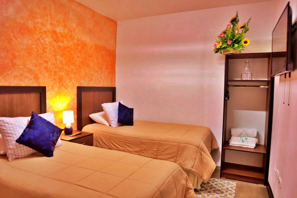 Habitación de hotel con dos camas, una mesita de noche con lámpara, un armario y una pequeña mesa con un jarrón de flores.