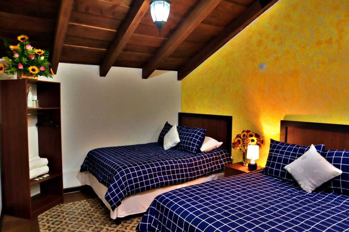Una habitación con dos camas, vigas de madera en el techo, una mesita de noche con una lámpara y una estantería con un jarrón de flores.