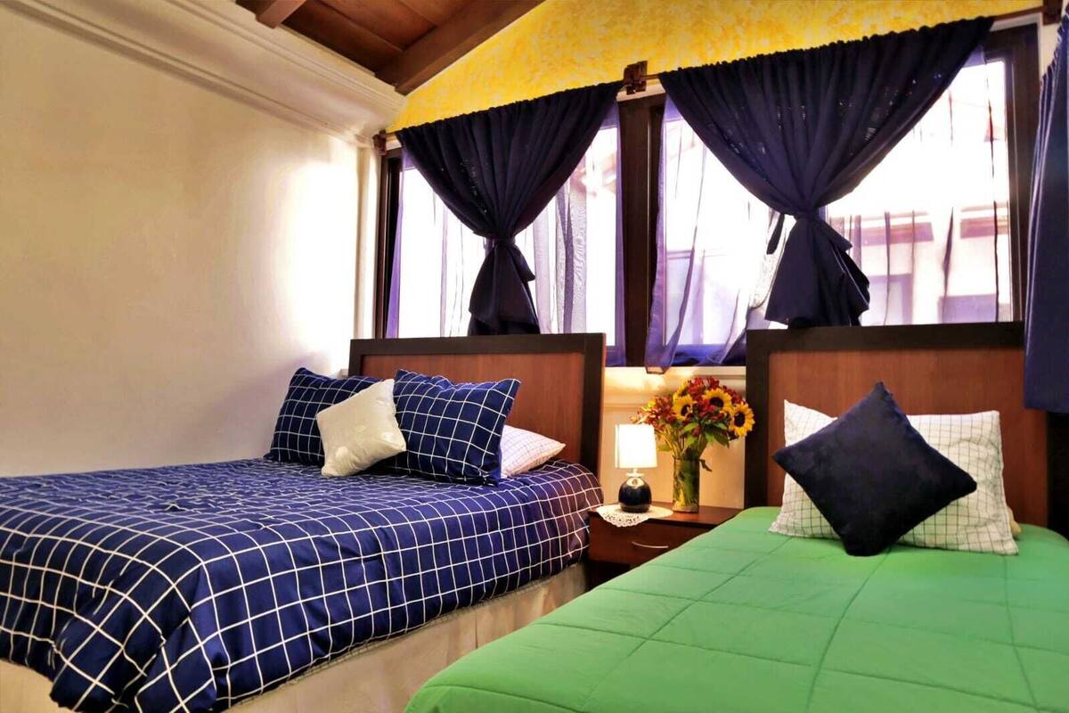 Habitación de hotel con dos camas, cada una con una colcha de diferente estampado. Entre las camas, una mesita de noche con una lámpara y un jarrón de flores.