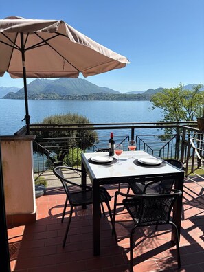 A terrace with a table and chairs, a bottle of wine, and a glass of red wine, overlooking a lake with mountains in the background.