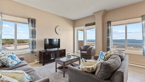 A living room with a TV, sofa, armchair, and a view of the sea. - Seawatch Oceanfront Unit 602 (Old Orchard Beach)