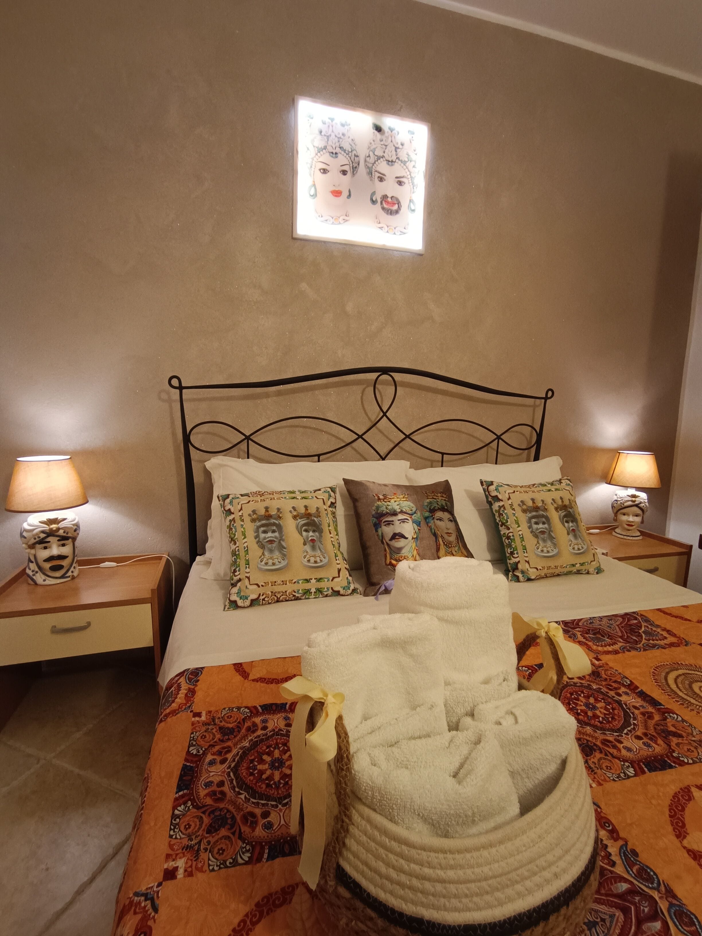 Un lit bien fait, avec une literie à motifs, deux coussins décoratifs, une lampe fixée au mur et une lampe de chevet posée sur une table de nuit en bois.
