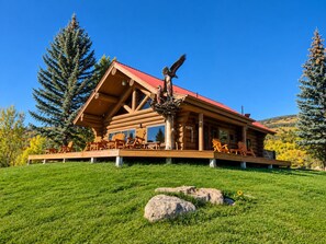 A log cabin with a red roof, wooden deck with chairs, and a bird sculpture.