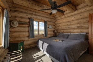 Une chambre dans un chalet en rondins, avec un lit, un ventilateur de plafond, une fenêtre avec des rideaux et un panneau accroché au mur.