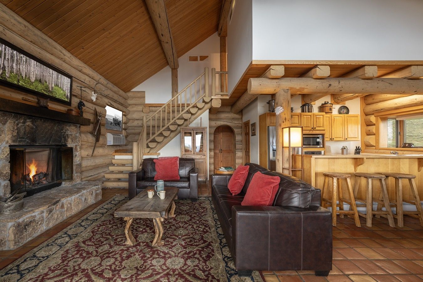 A log cabin interior with a stone fireplace, leather sofa, wooden staircase, and a kitchen area.
