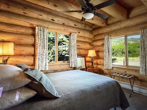 Une chambre à coucher située dans une cabane en rondins, avec un lit, deux fenêtres, un ventilateur de plafond et une table de chevet avec une lampe.