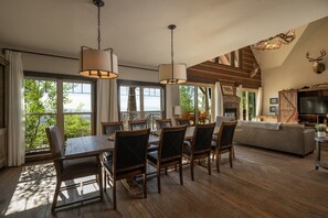 A spacious living area with a dining table, chairs, a sofa, and a fireplace. There are large windows offering a view of the outdoors.