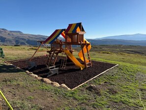 Un terrain de jeux avec un toboggan et une structure d’escalade, situé dans un champ herbeux avec des montagnes en arrière-plan.