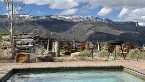 Une piscine rectangulaire bordée de briques, entourée de chaises en bois et d’une table en bois rustique.