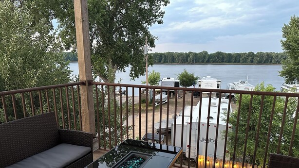 Un balcon doté d’une table à plateau en verre, de meubles en osier et offrant une vue sur un lac, avec des camping-cars visibles au loin.