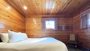A wooden cabin bedroom with a bed, a chair, and a window with curtains.