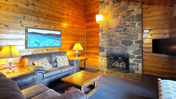 A cozy cabin interior with a stone fireplace, leather sofa, wooden walls, and a mounted television.