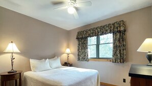 Ein ordentlich eingerichtetes Schlafzimmer mit einem großen Bett, zwei Nachttischen mit Lampen, einem Deckenventilator, einem Fenster mit Vorhängen und einer hölzernen Kommode.