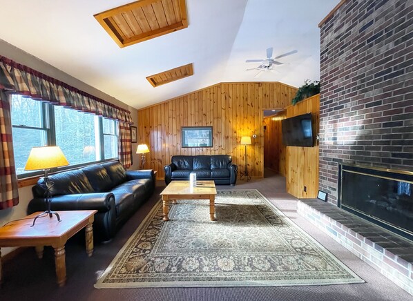 A cozy living room with wooden walls, a fireplace, a black leather sofa, a coffee table, and a patterned rug. - Charming Lake Front Cabin in the Poconos (Stroudsburg)