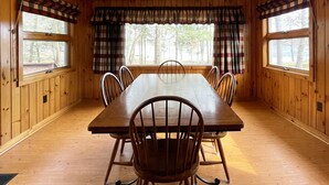 Ein Zimmer mit Holzwänden, einem großen Esstisch mit Stühlen, einem Deckenventilator und großen Fenstern mit Karottenvorhängen.