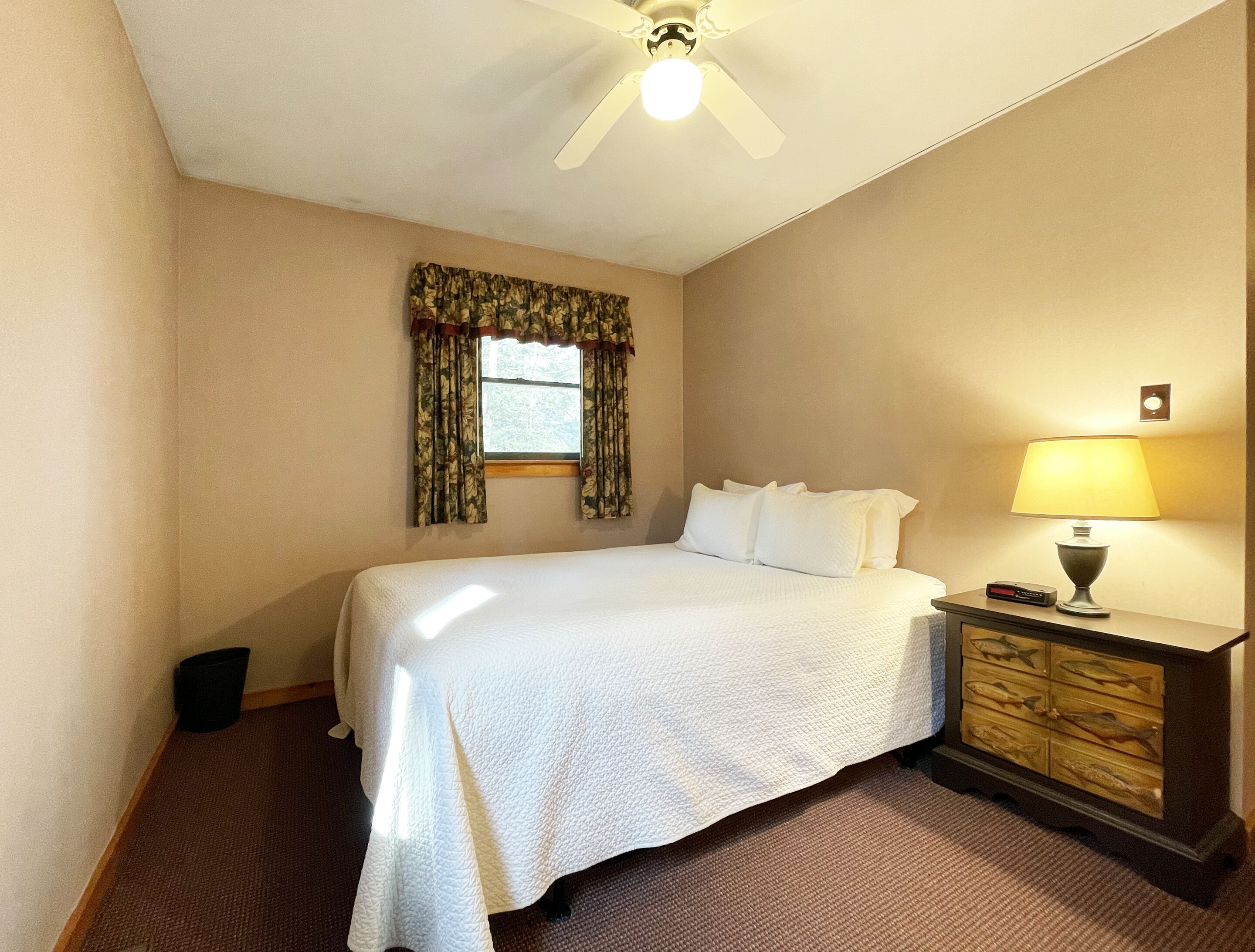 A hotel room with a bed, a nightstand, a lamp, a window with curtains, and a ceiling fan.