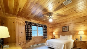 Un dormitorio de cabaña de madera con una cama, dos mesitas de noche con lámparas, una ventana con cortinas, un ventilador de techo y un cuadro colgado en la pared.