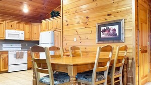 Interior de una cabaña de madera con una mesa de comedor, sillas, una cocina equipada y un cuadro en la pared.