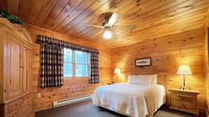 A wooden cabin bedroom with a bed, two bedside lamps, a ceiling fan, and a window with curtains. - Luxurious Lake View Log Cabin with Fire Pit in the Poconos (Stroudsburg)