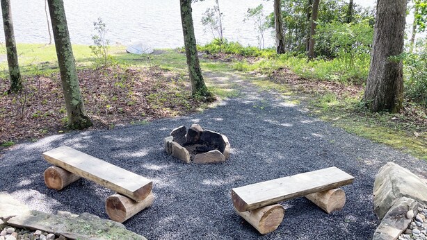A serene outdoor seating area with two wooden benches and a fire pit surrounded by gravel, overlooking a lake. - Luxurious Lake View Log Cabin with Fire Pit in the Poconos (Stroudsburg)