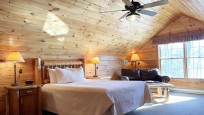 Un dormitorio rústico de cabaña con una cama, dos mesitas de noche con lámparas, un ventilador de techo y un ventanal con vistas a árboles.