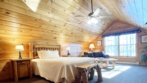 A cozy log cabin bedroom with a wooden ceiling, a bed with a headboard, two bedside tables with lamps, a bench, and a large window with a view of trees. - Luxurious Lake View Log Cabin with Firepit in the Poconos (Stroudsburg)
