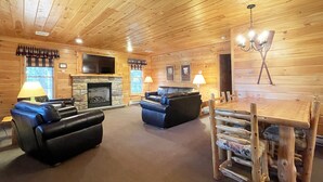 A log cabin interior with a fireplace, leather sofas, a dining table, and a chandelier. - Luxurious Lake View Log Cabin with Firepit in the Poconos (Stroudsburg)