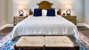 Una cama bien hecha con cabecera de madera, dos mesitas de noche con lámparas, y un banco con cojín estampado.