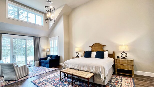 Un dormitorio amplio con una cama grande, un banco, un sillón azul y una mesita de noche con lámpara.