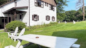 A white building with brown shutters, a covered porch, and a picnic table with chairs in a grassy area. - Poconos Rustic 1BR in the Poconos on Private Resort (Stroudsburg)