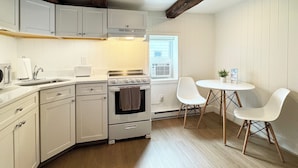 A kitchen with white cabinets, a stove, and a sink. There is a window with an air conditioner, and a small table with two chairs. - Poconos Rustic 1BR in the Poconos on Private Resort (Stroudsburg)