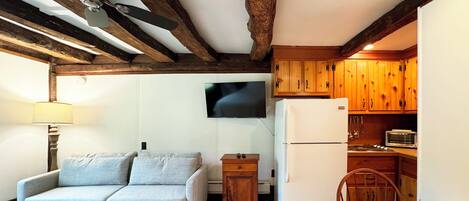 A cozy living room with a sofa, a small table, and a ceiling fan. There is a kitchen area with wooden cabinets and a refrigerator.